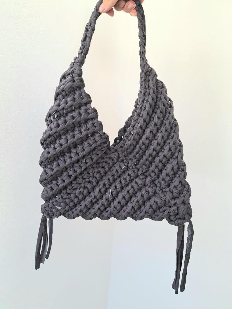 May include: A dark gray crochet halter top crafted from thick fabric yarn. The garment features a triangular bodice with a braided neck strap and two dangling fabric fringe accents at the bottom corners, showcasing a textured, chunky knit pattern.