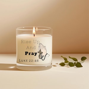 Puede incluir: Una vela encendida en un tarro de cristal transparente. La vela tiene un interior de cera blanca y presenta el texto "Rise Up And Pray" con una ilustración de manos rezando y una cruz. El texto "LUKE 22:46" también está impreso en el tarro.