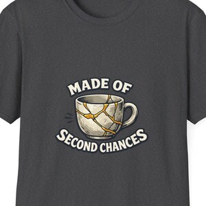 Puede incluir: Una camiseta gris carbón oscuro con un gráfico de una taza de cerámica blanca con grietas doradas, al estilo kintsugi. El texto "MADE OF SECOND CHANCES" aparece encima y debajo de la taza en una tipografía sans-serif de color crema.