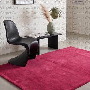 May include: A rectangular, solid magenta area rug with a plush, soft texture. The rug is positioned on a smooth, light gray floor in front of a black, curved chair and a black rectangular side table holding a vase with dried decorative foliage.