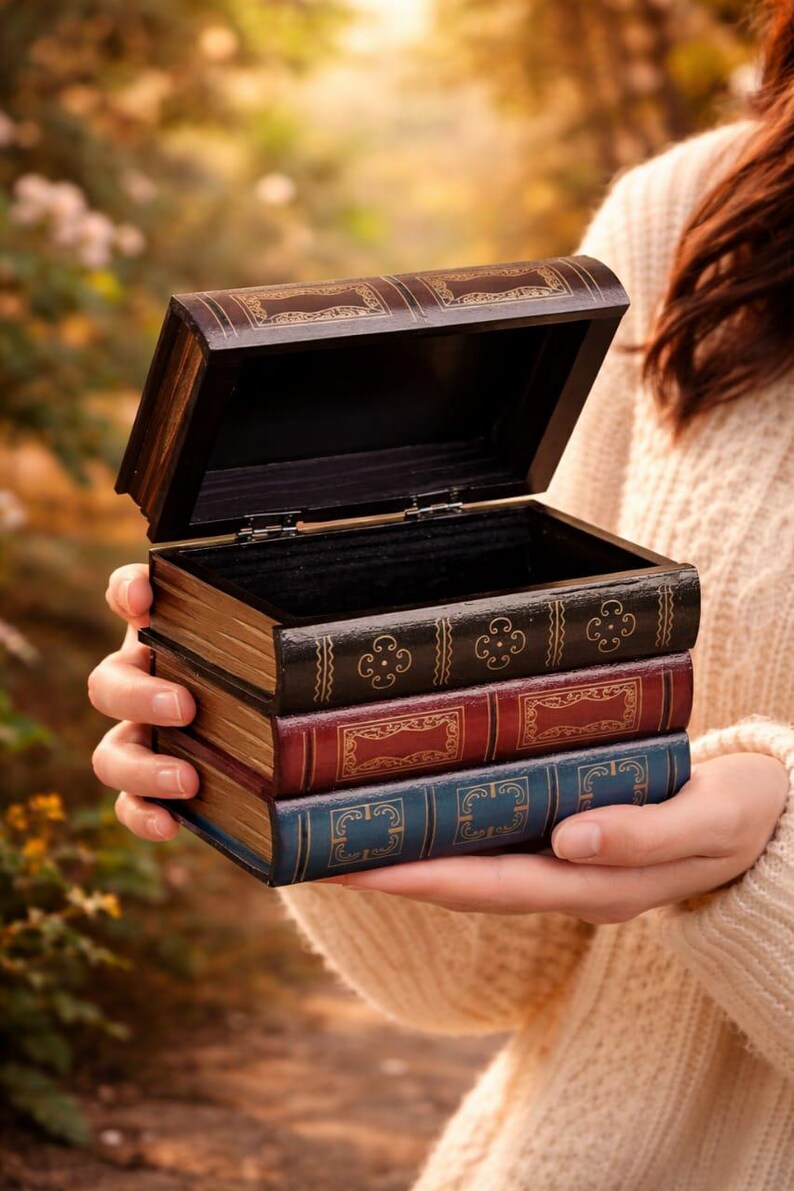 May include: A wooden box shaped like a stack of books, with the top book open. The box is dark brown with gold accents and the "book" spines are black, red, and blue. The box is held in a person's hands.