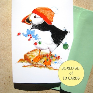 May include: A Christmas card featuring a cartoon puffin wearing a red and white Santa hat and holding a candy cane. The puffin is standing on a rock with Christmas lights and ornaments around it. The card says "Boxed Set of 10 Cards".