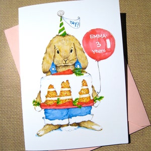 May include: A birthday card featuring a watercolor illustration of a brown bunny wearing a blue shirt and a party hat. The bunny is holding two carrot cakes. The card has the text "Yay!" and "Emma 3 years" on it.