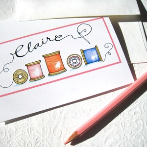 May include: A white card with pink trim and the name "Claire" written in cursive. There are five spools of thread in different colors, including red, orange, pink, and blue, with gold spools. The card is on a white surface.