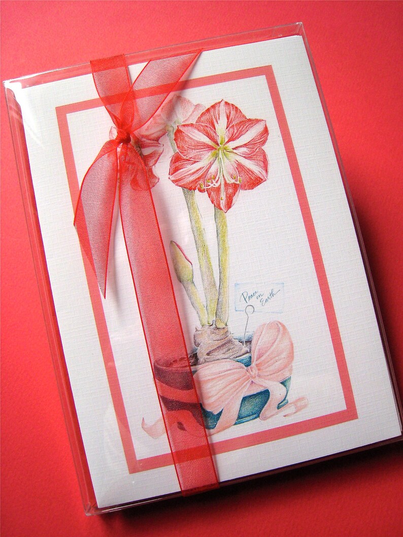 May include: A white card with a red border and a red ribbon tied around it. The card features a watercolor illustration of a red amaryllis flower in a pot with a pink bow. The text "Peace on Earth" is written on a small tag attached to the flower.