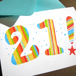 May include: A white card with the number 21 written in colorful stripes. The number 1 has an exclamation point next to it, also in colorful stripes. The card has a blue envelope on top of it.