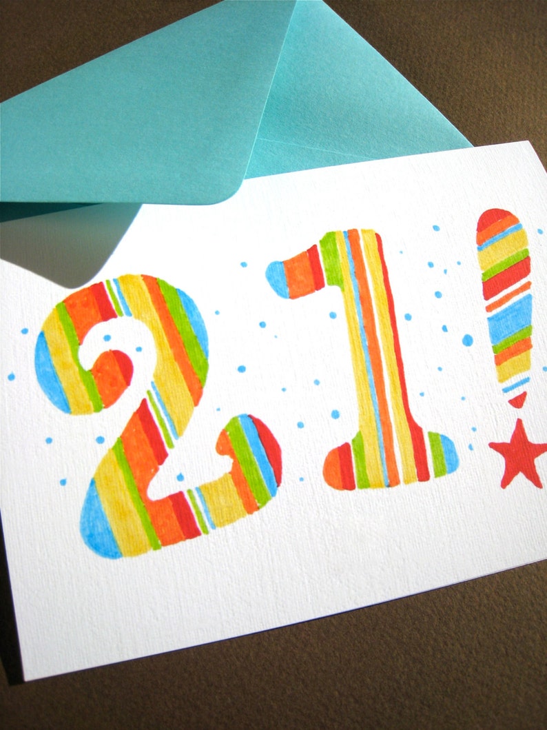 May include: A white card with the number 21 written in colorful stripes. The number 1 has an exclamation point next to it, also in colorful stripes. The card has a blue envelope on top of it.