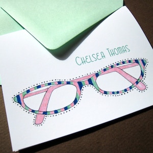 May include: A white card with the name "Chelsea Thomas" written in green text. A pink and blue striped eyeglass frame with black dots around the edge is drawn on the card. A small pink heart is drawn in the top right corner.