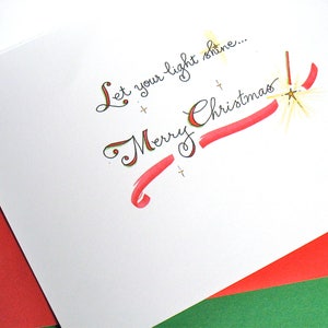 May include: A white card with red and gold accents. The card has the text "Let your light shine... Merry Christmas" written in cursive.