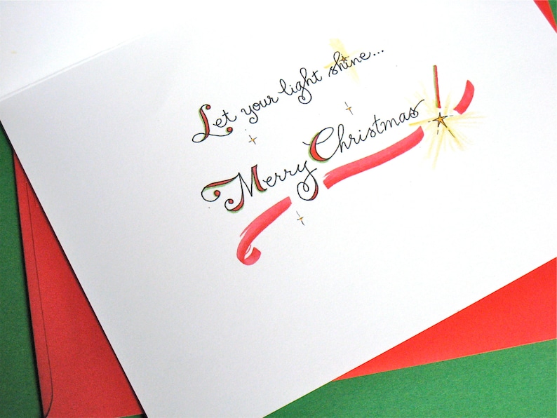 May include: A white card with red and gold accents. The card has the text "Let your light shine... Merry Christmas" written in cursive.