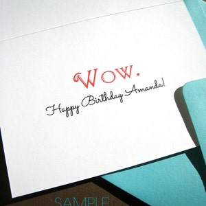 May include: A white card with the text "WOW." in red and "Happy Birthday Amanda!" in black script. The card is folded in half and a light blue envelope is partially visible in the bottom right corner. The text "SAMPLE Custom Name" is printed in black on the bottom of the card.