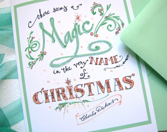 May include: A Christmas card with the text "there seems a Magic in the very NAME of CHRISTMAS" in green and orange lettering. The card has a green border and is accompanied by a green envelope. The design includes decorative flourishes and the signature "Charles Dickens".
