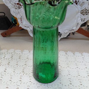 May include: A tall, green glass vase with a flared, ruffled top. The vase has a cylindrical body with vertical textured lines. The rim of the vase has a dark, possibly black, trim. The vase is sitting on a white, lace-like surface.