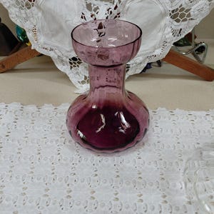 May include: A vintage, amethyst-colored glass vase with a bulbous base and a flared rim. The vase has a textured surface and is sitting on a white, eyelet-style tablecloth. The background includes a white lace doily and a wooden drying rack.