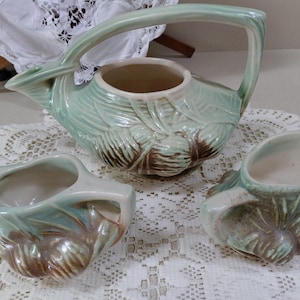 May include: A vintage ceramic tea set in a soft green and brown color scheme. The set includes a teapot with a handle and two smaller serving pieces, all with a textured, leaf-like design. The set is displayed on a white lace tablecloth.