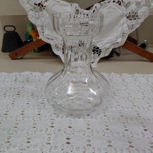 May include: A clear, textured glass vase with a bulbous base and a flared top. The vase has a honeycomb-like pattern on the lower portion and a ribbed design on the neck. It sits on a white, patterned tablecloth.