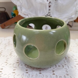 May include: A round, light green ceramic candle holder with three large circular openings. The candle holder has a glossy finish and sits on a white, lace-like surface. The openings allow light to shine through, creating a decorative effect.