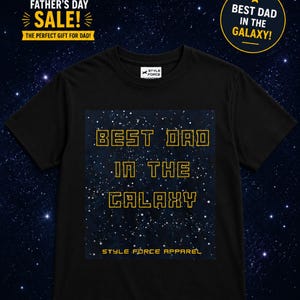 May include: A black short-sleeved t-shirt featuring a rectangular graphic of a starry night sky. The text "BEST DAD IN THE GALAXY" is printed in a yellow, blocky, outlined font, with "STYLE FORCE APPAREL" written in smaller yellow text below.