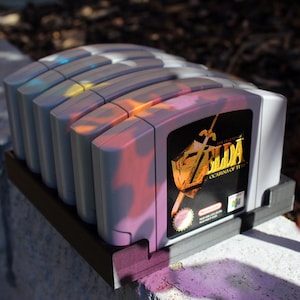 May include: A row of five grey Nintendo 64 video game cartridges held in a black plastic stand. The front cartridge features a label with the gold text The Legend of Zelda Ocarina of Time, a sword and shield graphic, and the Nintendo 64 logo.