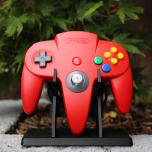 May include: A red Nintendo 64 video game controller featuring a grey directional pad, a central grey analogue stick with a white top, a red start button, and four coloured action buttons in yellow, green, and blue, mounted on a black display stand.