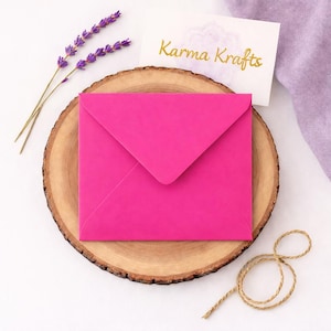 May include: A vibrant pink envelope on a wooden slice, with lavender sprigs and a card bearing the gold text "Karma Krafts". A length of twine is also present.
