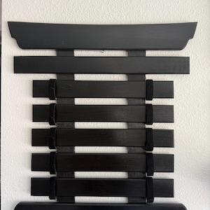 May include: A black wooden wall-mounted display rack featuring a series of horizontal slats connected by vertical black fabric straps. The structure has a wider top and bottom piece with curved edges, providing a grid-like frame for hanging items.