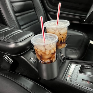May include: Two iced McCafe drinks with red and white striped straws in a classic car's cup holder. The drinks are in clear plastic cups, filled with ice and a dark beverage. The car's interior is black leather.