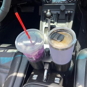 May include: A black dual cup holder accessory installed in a vehicle centre console. It holds a clear plastic cup with a purple beverage and a red straw, alongside a light purple insulated tumbler with a black lid containing a coffee drink.