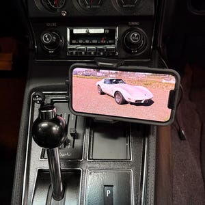 Może przedstawiać: Wnętrze klasycznego samochodu z deską rozdzielczą, wskaźnikami, radiem i dźwignią zmiany biegów. Smartfon wyświetla biały samochód sportowy. Deska rozdzielcza jest czarna ze srebrnymi akcentami.