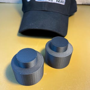 Pode incluir: Boné de beisebol preto com o texto "Corvette Sting Ray" em branco. Dois objetos cilíndricos cinza escuro com um cilindro menor no topo. Fundo amarelo-alaranjado.