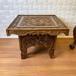 May include: A square, wooden side table with intricate floral carvings. The table has a dark brown finish and features detailed relief patterns on the tabletop and legs. The design is ornate and traditional, suitable for a living room or bedroom.