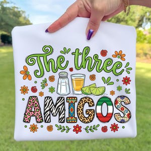 Puede incluir: Camiseta blanca con la frase "The three AMIGOS" en verde, con elementos decorativos coloridos. El diseño incluye un salero, un chupito de tequila, rodajas de lima y la palabra AMIGOS en varios patrones y colores.