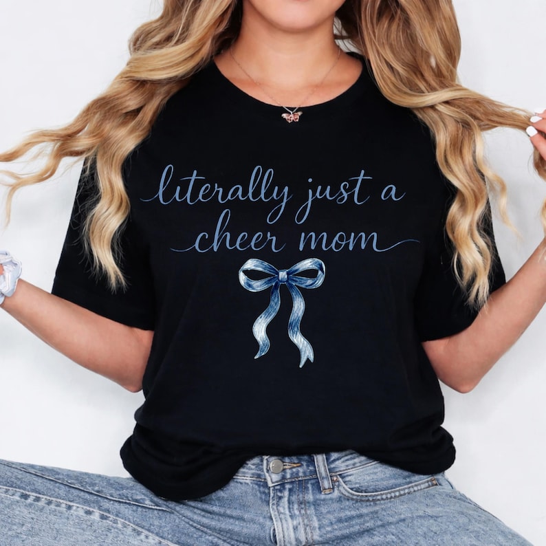 May include: Black t-shirt with the text "literally just a cheer mom" in a light blue script font. A blue ribbon graphic is below the text. The shirt is a crew neck style.