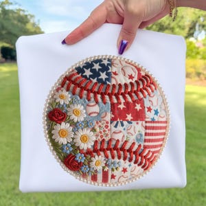 Peut inclure: Sac fourre-tout blanc avec un motif de baseball. Le baseball est orné de coutures rouges, de broderies florales et de motifs d'étoiles, de rayures et de balles de baseball. Une bordure de perles entoure le motif de baseball.