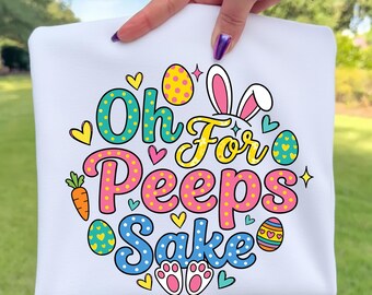 Könnte beinhalten: Weißes T-Shirt mit dem Schriftzug "Oh For Peeps Sake" in bunten Buchstaben. Das Design umfasst Ostereier, Hasenohren, Herzen, eine Karotte und Hasenpfoten. Das T-Shirt wird von einer Hand gehalten.