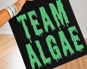 Könnte beinhalten: Ein schwarzer Stoffartikel mit dem Text "TEAM ALGAE", der in einer hellgrünen, tropfenden, schleimartigen Schrift gedruckt ist. Die Buchstaben haben ein geschmolzenes, flüssiges Aussehen und bilden ein kontrastreiches Grafikdesign.