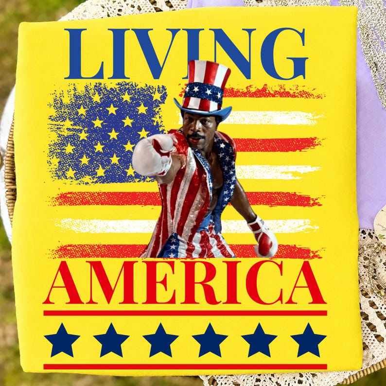 May include: A bright yellow t-shirt featuring the text LIVING AMERICA in blue and red. The graphic shows a person wearing a red, white, and blue top hat and boxing attire, posed in front of a stylised American flag with blue stars at the bottom.