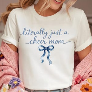 May include: White t-shirt with the text "literally just a cheer mom" in blue script. A blue watercolor bow is below the text. The shirt has short sleeves and is made of a soft material.