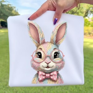 May include: White fabric with a colorful bunny graphic. The bunny has pink glasses, a pink bow tie with white polka dots, and a multi-colored fur pattern. The background is a blurred green lawn.