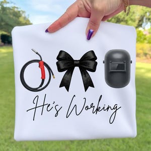 May include: White sweatshirt with a black bow, welding equipment graphic, and a welding helmet. The text "He's Working" is written in a cursive font. The sweatshirt is held up by a hand with purple nail polish.