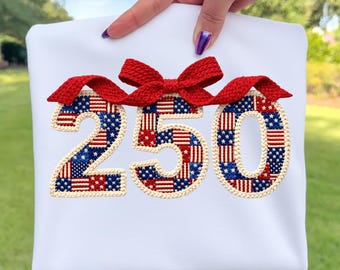 Könnte beinhalten: Weißes T-Shirt mit der Zahl "250", die in Rot, Weiß und Blau gestickt ist und an die amerikanische Flagge erinnert. Oben ist eine rote Schleife angebracht. Das Design ist mittig auf dem Shirt platziert.