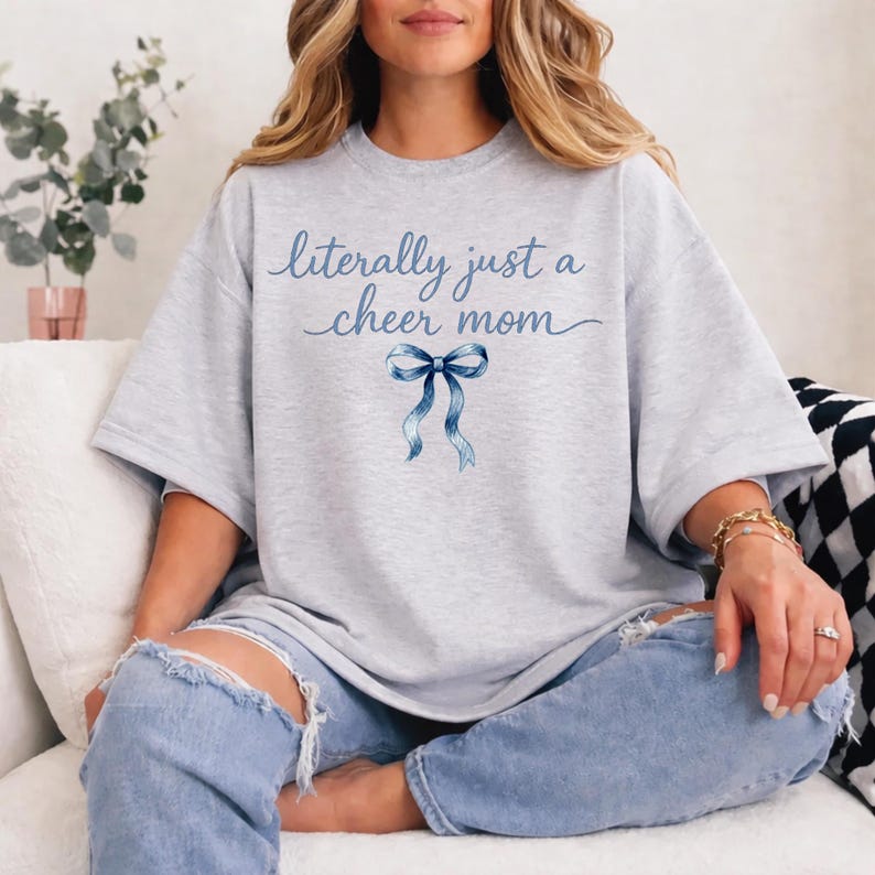 May include: A light gray t-shirt with the text "literally just a cheer mom" in blue script. A blue bow graphic is centered below the text. The shirt is paired with light blue jeans.