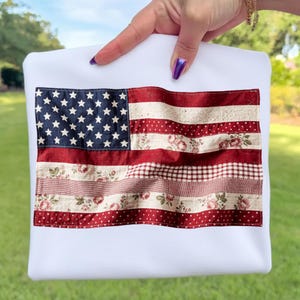 Op de afbeelding: Witte draagtas met een patchwork ontwerp van de Amerikaanse vlag. De vlag heeft een blauw sterrenveld en rood-witte strepen, met bloemen-, stippen- en ruitpatronen. De tas wordt door een hand omhoog gehouden.