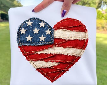 Könnte beinhalten: Weißes T-Shirt mit einem herzförmigen Design, das die amerikanische Flagge darstellt. Das Herz ist in zwei Teile geteilt: ein blaues Denim-Patch mit weißen Sternen und ein rot-weiß gestreifter Bereich. Das Design besteht aus strukturiertem Stoff.
