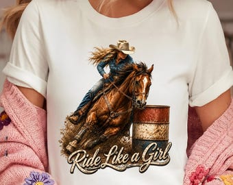 Könnte beinhalten: Weißes T-Shirt mit einer Grafik einer Frau auf einem braunen Pferd, das um ein Fass reitet. Die Frau trägt einen Cowboyhut und ein Jeanshemd. Das Fass hat rot-weiß-blaue Streifen. Der Text "Ride Like a Girl" steht darunter.