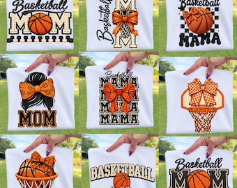 Könnte beinhalten: Weiße Sweatshirts mit Basketball-Motiven. Die Designs umfassen Basketbälle, Schleifen und die Wörter "Basketball Mom" und "Basketball Mama" in verschiedenen Schriftarten. Die Sweatshirts werden von Händen gehalten.