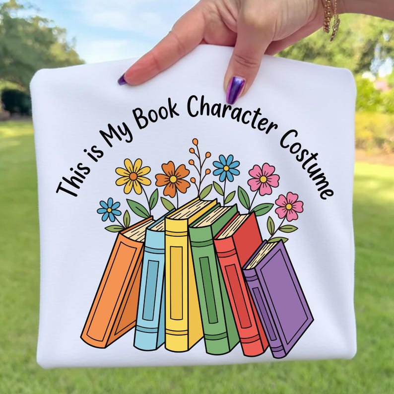 May include: White sweatshirt with the text "This is My Book Character Costume" in black. The design features a stack of colorful books with flowers. The books have orange, blue, yellow, green, red, and purple spines.
