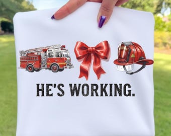 Könnte beinhalten: Weißes Sweatshirt mit einem roten Feuerwehrauto, einer roten Schleife und einem roten Feuerwehrhelm. Der Text "HE'S WORKING." ist darunter in Schwarz gedruckt.