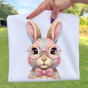 Könnte beinhalten: Weißer Stoff mit einer bunten Hasenillustration. Der Hase trägt eine rosa Brille und eine Fliege, mit mehrfarbigem Fell in Rosa, Blau und Beige. Das Bild wird von einer Hand vor einem grünen Hintergrund gehalten.