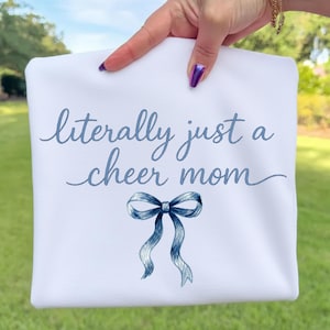 May include: White t-shirt with the text "literally just a cheer mom" in blue script. A blue bow graphic is below the text. The shirt is held up by a hand with purple nail polish, against a green background.